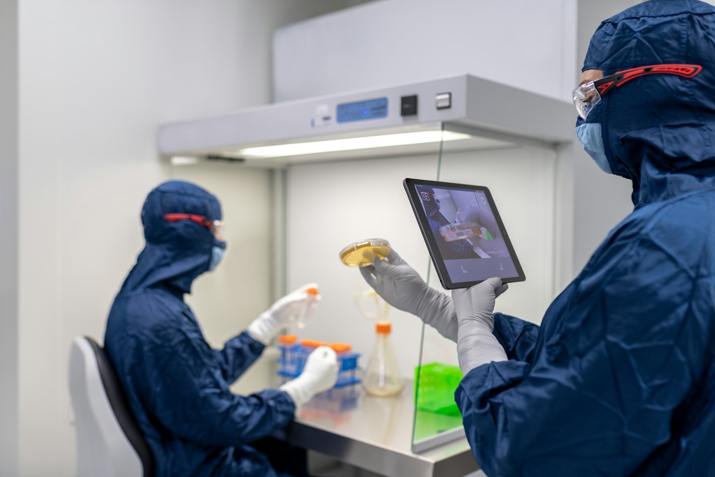 Scientists in hazmat suits use a tablet and test tubes in a lab.